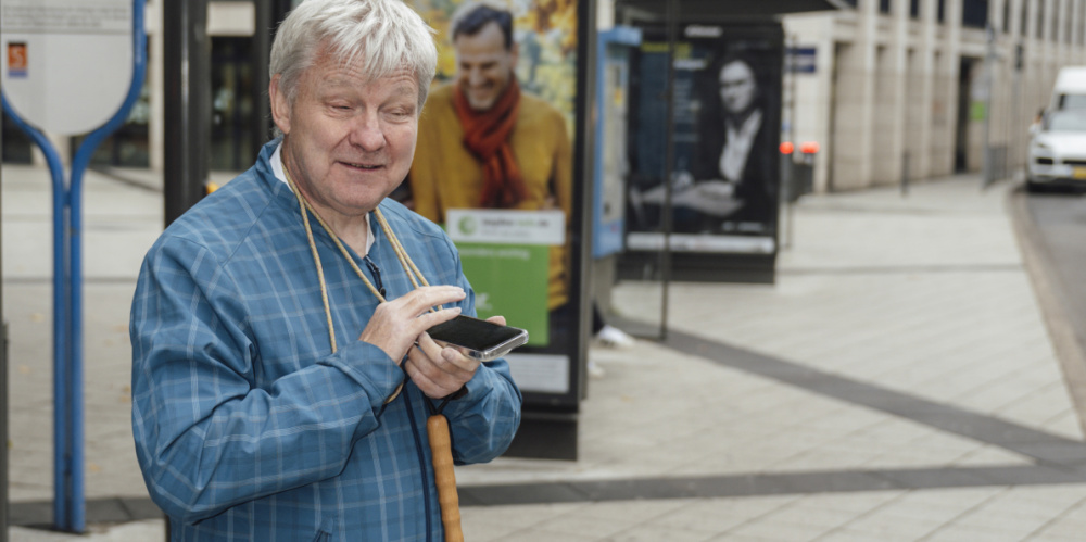 Ein älterer Mann mit einem Blindenstock nutzt an einer Haltestelle das Barrierefreie Informationssystem auf seinem Handy.